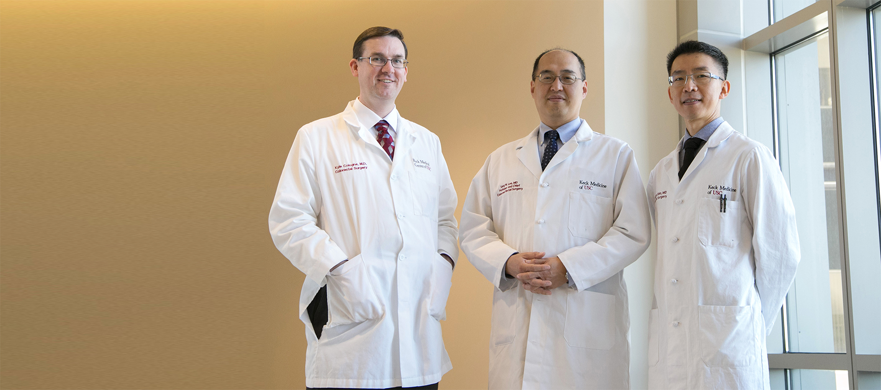 Three of Keck Medicine's colorectal surgeons, who specialize in colon and anal cancer, stand by a tan wall and a large window