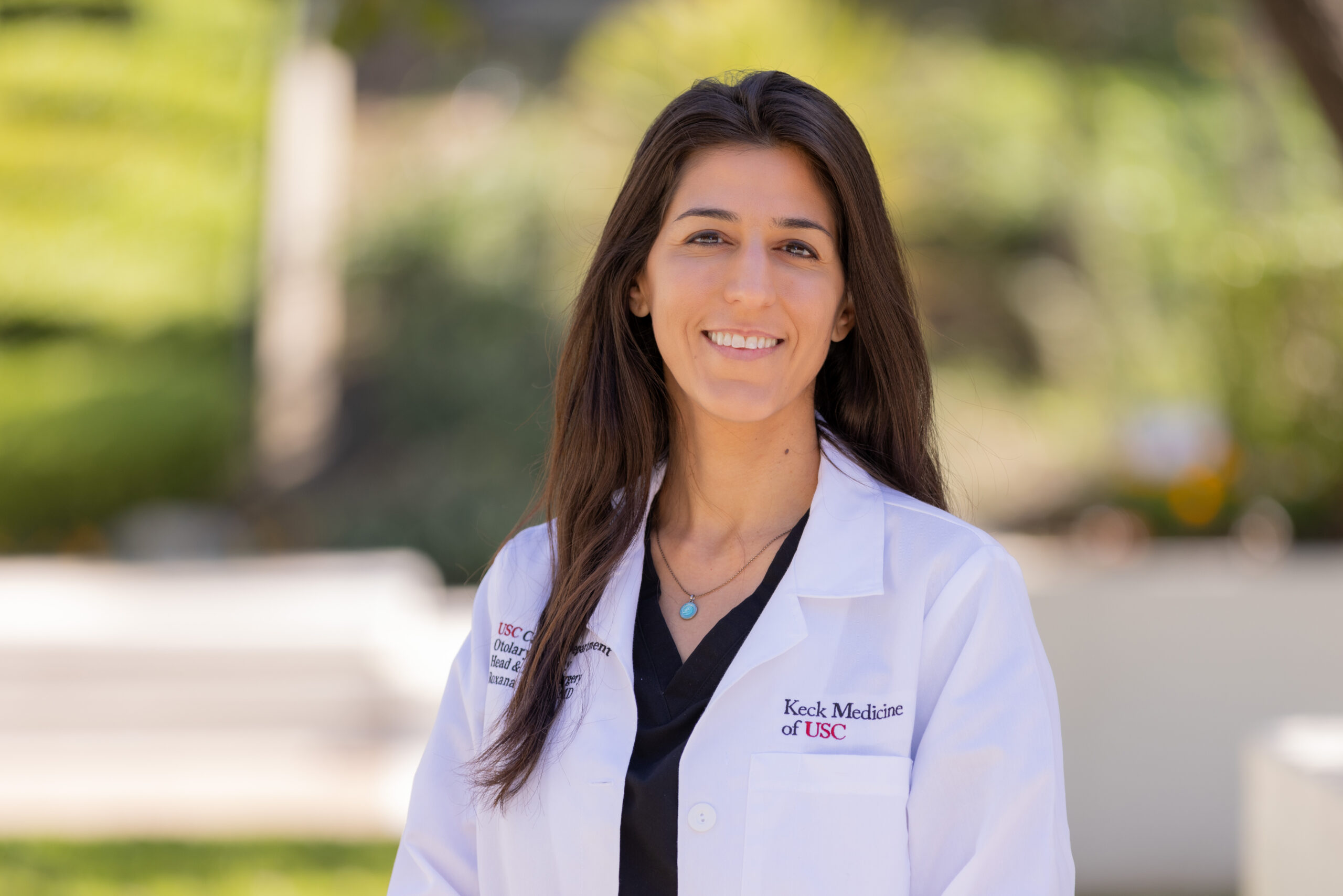 An outdoor portrait Dr. Roxana Moayer smiling in a white medical coat.